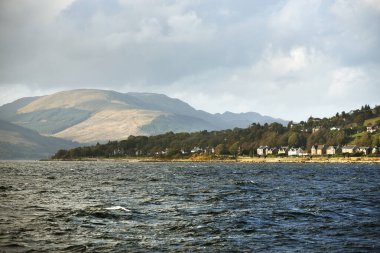 Sudan kayalık nehir kıyılarının panoramik görüntüsü. Arka planda kır evleri, ağaçlar, tepeler ve dağlar var. Bulutlu mavi gökyüzü. Gare Loch, Clyde Firth, İskoçya, İngiltere