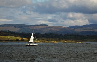 Bulutlu bir günde, yakın planda, Crinan Kanalı 'nda siyah şalopeli yat gezintisi. Arka planda kayalık kıyılar var. Dramatik mavi gökyüzü. Ardrishaig, İskoçya, İngiltere. Spor ve eğlence teması