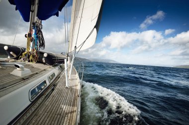 Bulutlu bir günde ahşap meşe güverteli, yelkenli, modern bir yat. Direğe ve yelkenlere bir manzara. Sudan kayalık nehir kıyılarının panoramik görüntüsü. Arka planda kır evleri, ağaçlar, tepeler ve dağlar var. Gare Gölü, Clyde 'ın Firth' i..