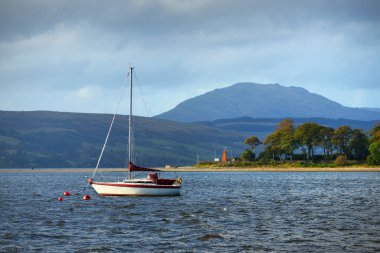 Güneşli bir günde yelkenli dolu bir yat. Arkasında tepeler, ormanlar ve kır evleri olan kayalık kıyılar. Rhu marinası, Clyde Firth, İskoçya, İngiltere