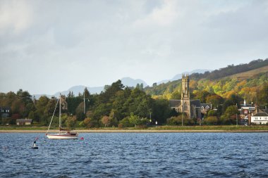 Güneşli bir günde yelkenli dolu bir yat. Arkasında tepeler, ormanlar ve kır evleri olan kayalık kıyılar. Rhu marinası, Clyde Firth, İskoçya, İngiltere