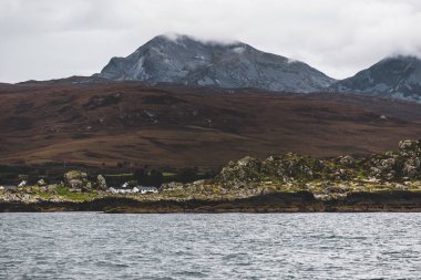Bulutlu mavi gökyüzünün altında Jura Paps (Beinn an ir) 'ın en yüksek dağ zirvesi yakınındaki kayalık kıyısı. Yelkenliden panoramik manzara. Jura Adası, Inner Hebrides, İskoçya, İngiltere.