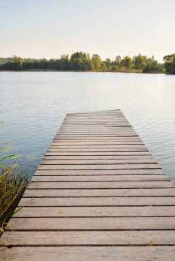 Gün batımında göl kıyısında. Ahşap iskeleye yakın çekim. Arka planda orman ve köy var. Sudaki yansımalar. Idyllic kırsal kesim. Pastoral manzara. Ekolojik tatil köyü, balık tutma, yüzme. Sonbaharın başı