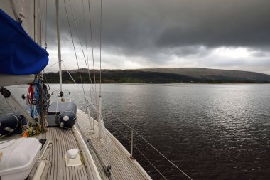Karanlık fırtına gökyüzü. Bulutlu bir günde ahşap meşe güverteli, yelkenli, modern bir yat. Güverteden pruvaya bir manzara. Bute 'un kayalık kıyılarının sudan panoramik görüntüsü. Tepeler ve dağlar arka planda. Bute Adası, Firth of