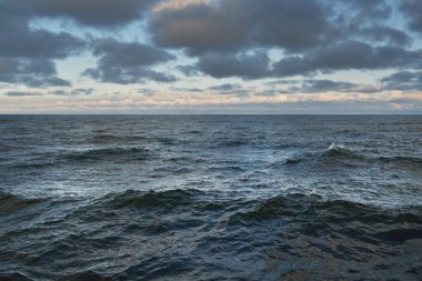 Gün batımında açık Baltık denizinin panoramik görüntüsü. Renkli parlak kümülüs bulutlarıyla dramatik bir gökyüzü. Su yüzeyi dokusu yakın çekim. Dengesiz hava, kış, iklim değişikliği, okyanus kabarması, doğa.