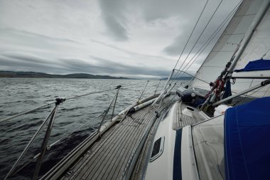 Bulutlu bir günde ahşap meşe güverteli, yelkenli, modern bir yat. Direğe ve yelkenlere bir manzara. Su, yakından sıçrıyor. Tarbert, İskoçya, İngiltere