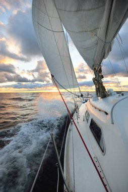 Gün batımında açık denizde yelken açmak. Güvertenin yakın görüntüsü, direk ve yelkenler. Yağmurdan sonra açık gökyüzü, çarpıcı parlayan bulutlar, altın güneş ışığı, dalgalar ve su sıçramaları, kasırga. Destansı deniz manzarası