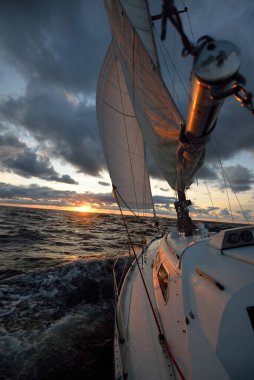 Gün batımında açık denizde yelken açmak. Güvertenin yakın görüntüsü, direk ve yelkenler. Yağmurdan sonra açık gökyüzü, çarpıcı parlayan bulutlar, altın güneş ışığı, dalgalar ve su sıçramaları, kasırga. Destansı deniz manzarası