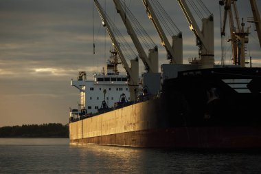 Gün batımında limana demir atmış büyük bir kargo gemisi. Vinçler arka planda. Riga, Baltık Denizi, Letonya. Yük taşımacılığı, küresel iletişim, lojistik, çevresel hasar