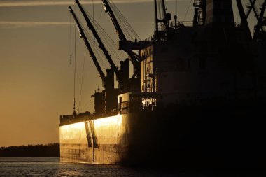 Gün batımında limana demir atmış büyük bir kargo gemisi. Vinçler arka planda. Riga, Baltık Denizi, Letonya. Yük taşımacılığı, küresel iletişim, lojistik, çevresel hasar