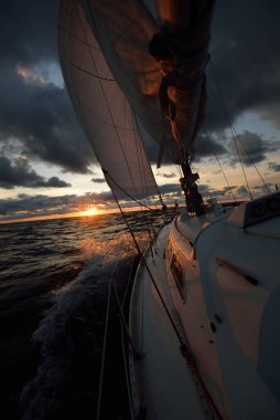 Gün batımında açık denizde yelken açmak. Güvertenin yakın görüntüsü, direk ve yelkenler. Yağmurdan sonra açık gökyüzü, çarpıcı parlayan bulutlar, altın güneş ışığı, dalgalar ve su sıçramaları, kasırga. Destansı deniz manzarası