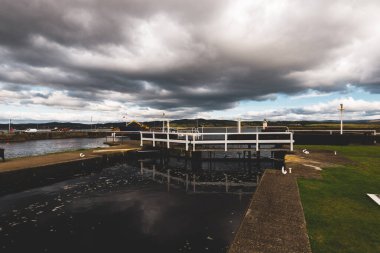 Küçük bir liman ve eski deniz fenerine kadar boş bir gezinti alanı, yakın plan. Arka planda kayalık kıyılar var. Dramatik bulut manzarası. Ardrishaig, Crinan Kanalı, İskoçya, İngiltere. Seyahat yerleri, gezi teması