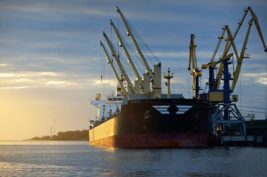 Gün batımında limana demir atmış büyük bir kargo gemisi. Vinçler arka planda. Riga, Baltık Denizi, Letonya. Yük taşımacılığı, küresel iletişim, lojistik, çevresel hasar