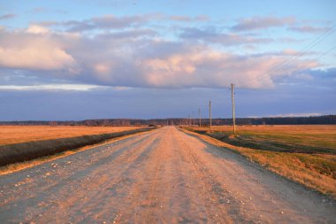 Gün batımında Letonya 'da tarladan geçen boş bir köy yolu. Transformatör direkleri yakın çekim. Arabadan bir manzara. Idyllic kırsal kesim. Renkli parlayan bulutlar. Ulaşım, yolculuk, özgürlük kavramları