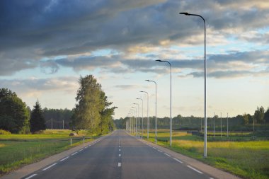 Gün batımında yeşil kırlarda boş bir otoyol (yeni asfalt yol). Sokak lambaları yakın çekim. Dramatik parlayan bulutlar. Idyllic kırsal kesim. Seyahat yerleri, tatiller, sürüş, özgürlük