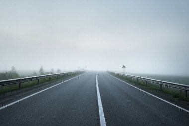 Gün doğumunda sisli bir havada boş otobanın tarlalar ve ormanlar arasındaki panoramik görüntüsü. Avrupa. Ulaşım, lojistik, seyahat, yolculuk, özgürlük, sürüş. Kırsal alan