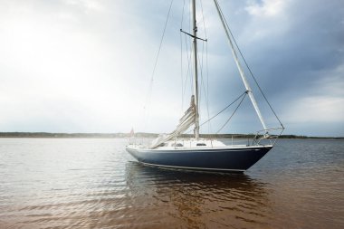 Kumlu kıyıya yakın sığ sularda demirli mavi şalopeli bir yat. Kara fırtına bulutları. Boş zaman aktivitesi, spor, eğlence teması. Glomma Nehri, Oslofjord, Norveç