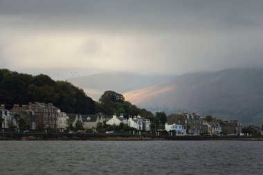 Fırtına gökyüzü. Küçük bir kasabanın panoramik manzarası sudan Rothesay. Köy evleri ve arabalar yakın plan. Arka planda ormanlar, tepeler ve dağlar. Bute Adası, Clyde Firth, İskoçya, İngiltere