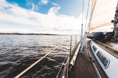 Yağmurdan sonra Tarbert 'in kayalık kıyılarında seyir halindeki ahşap meşe güvertesiyle donatılmış modern bir yat. İskoçya, İngiltere. Güverteden pruvaya, direğe ve yelkenlere yakın görüntü. Açık mavi gökyüzü