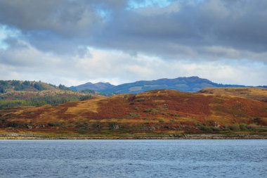 Sudan kayalık nehir kıyılarının panoramik görüntüsü. Arka planda ağaçlar, tepeler ve dağlar var. Bulutlu mavi gökyüzü. Gare Loch, Clyde Firth, İskoçya, İngiltere
