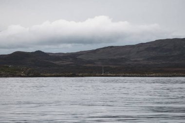 Bulutlu mavi gökyüzünün altındaki Jura Paps dağlarının yakınlarındaki kayalık sahilin panoramik manzarası. Yattan bir manzara. Jura Adası, Inner Hebrides, İskoçya, İngiltere. Seyahat yerleri, işaretler