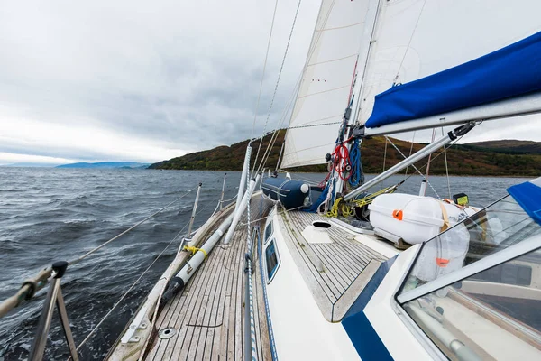 Sloop rigged modern yacht with wooden teak deck sailing near the rocky shores of Tarbert after the rain. Scotland, UK. Close-up view from the deck to the bow, mast and sails. Dramatic storm sky