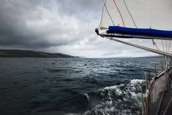 Storm sky. Sloop rigged modern yacht with wooden teak deck sailing  in the Firth of Clyde. Forests, hills and mountains of the Bute Island in the background. Scotland, UK