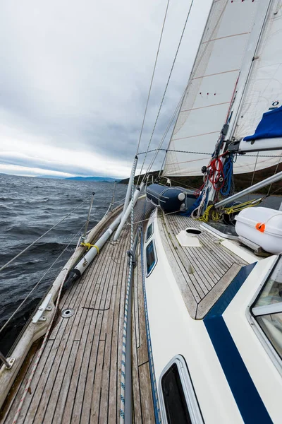 Sloop rigged modern yacht with wooden teak deck sailing near the rocky shores of Tarbert after the rain. Scotland, UK. Close-up view from the deck to the bow, mast and sails. Dramatic storm sky