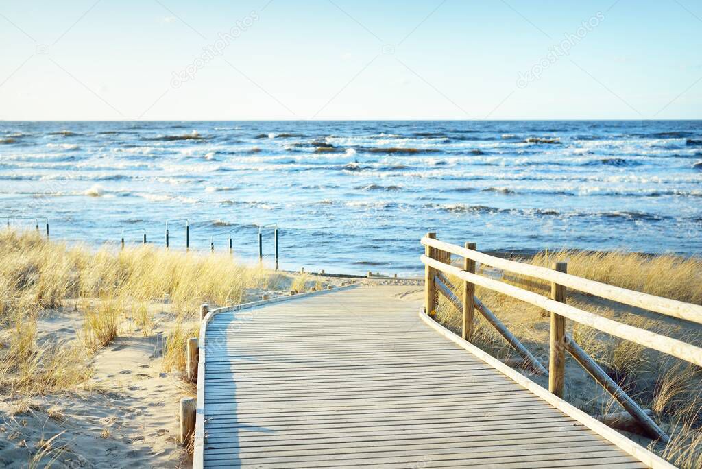 A wooden pathway to the Baltic sea at sunset. Sand dunes and plants in the background. Clear blue evening sky. Idyllic seascape. Latvia