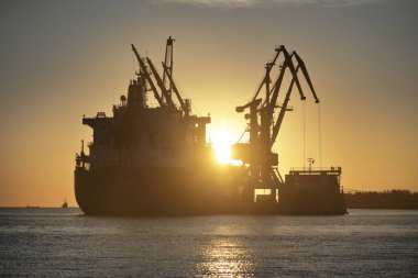 Gün batımında limana demir atmış büyük bir kargo gemisi. Vinçler arka planda. Riga, Baltık Denizi, Letonya. Yük taşımacılığı, küresel iletişim, lojistik, çevresel hasar