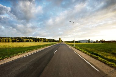 Gün batımında yeşil tarlalar ve huş ağacı ormanları boyunca boş bir otoyol (yeni asfalt yol). Sokak lambaları yakın çekim. Dramatik parlayan bulutlar. Seyahat yerleri, tatiller, sürüş, özgürlük