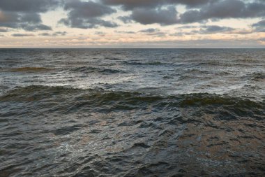 Gün batımında açık Baltık denizinin panoramik görüntüsü. Renkli parlak kümülüs bulutlarıyla dramatik bir gökyüzü. Su yüzeyi dokusu yakın çekim. Dengesiz hava, kış, iklim değişikliği, okyanus kabarması, doğa.