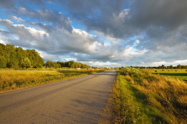 Gün batımında tarım arazisi ve ormandan geçen köy yolu. Dramatik bir gökyüzü. Arka planda saman yığınları var. Letonya 'da sonbaharın başlarında. Tatiller, seyahatler, araba kullanma, macera