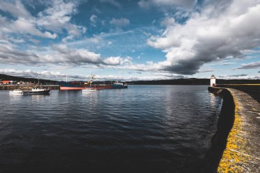 Küçük bir liman ve eski deniz fenerine kadar boş bir gezinti alanı, yakın plan. Arka planda kayalık kıyılar var. Dramatik bulut manzarası. Ardrishaig, Crinan Kanalı, İskoçya, İngiltere. Seyahat yerleri, gezi teması