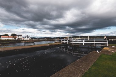 Küçük bir liman ve eski deniz fenerine kadar boş bir gezinti alanı, yakın plan. Arka planda kayalık kıyılar var. Dramatik bulut manzarası. Ardrishaig, Crinan Kanalı, İskoçya, İngiltere. Seyahat yerleri, gezi teması