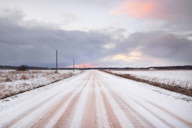 Tarlada karla kaplı kırsal yol. Elektrik hattı, trafo direkleri. Arabadan panoramik görüntü. Renkli bulutlar, dramatik günbatımı gökyüzü. Off-road, lojistik, kış lastikleri, uzak köy