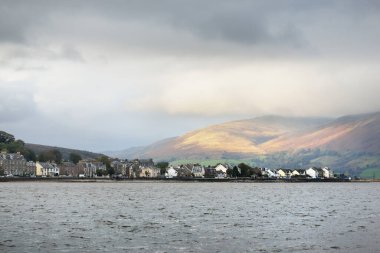 Fırtına gökyüzü. Küçük bir kasabanın panoramik manzarası sudan Rothesay. Köy evleri ve arabalar yakın plan. Arka planda ormanlar, tepeler ve dağlar. Bute Adası, Clyde Firth, İskoçya, İngiltere