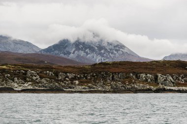 Bulutlu mavi gökyüzünün altında Jura Paps (Beinn an ir) 'in en yüksek dağ zirvesi yakınlarındaki kayalık sahilin panoramik görüntüsü. Yattan bir manzara. Jura Adası, Inner Hebrides, İskoçya, İngiltere.