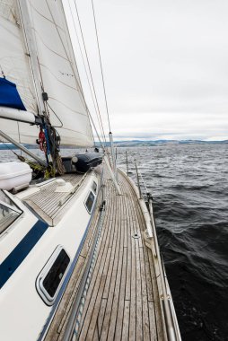 Yağmurdan sonra Tarbert 'in kayalık kıyılarında seyir halindeki ahşap meşe güvertesiyle donatılmış modern bir yat. İskoçya, İngiltere. Güverteden pruvaya, direğe ve yelkenlere yakın görüntü. Dramatik fırtına gökyüzü