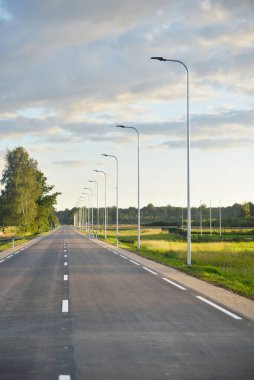 Gün batımında yeşil kırlarda boş bir otoyol (yeni asfalt yol). Sokak lambaları yakın çekim. Dramatik parlayan bulutlar. Idyllic kırsal kesim. Seyahat yerleri, tatiller, sürüş, özgürlük
