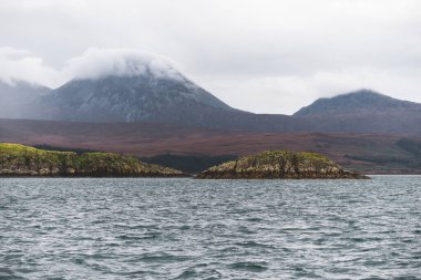 Bulutlu mavi gökyüzünün altında Jura Paps (Beinn an ir) 'ın en yüksek dağ zirvesi yakınındaki kayalık kıyısı. Yelkenliden panoramik manzara. Jura Adası, Inner Hebrides, İskoçya, İngiltere.
