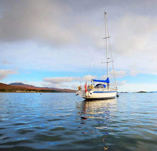 White sloop rigged yacht anchored on mooring near the shore of Craighouse at sunset. Mountains (Paps of Jura) in the background. Craighouse, Jura island, Inner Hebrides, Scotland, UK. Travel concepts