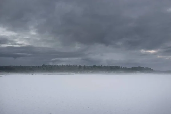 Snow-covered agricultural plowed field and forest under dramatic dark clouds before snowstorm. Winter rural scene. Nature, ecology, environment, climate change, global warming