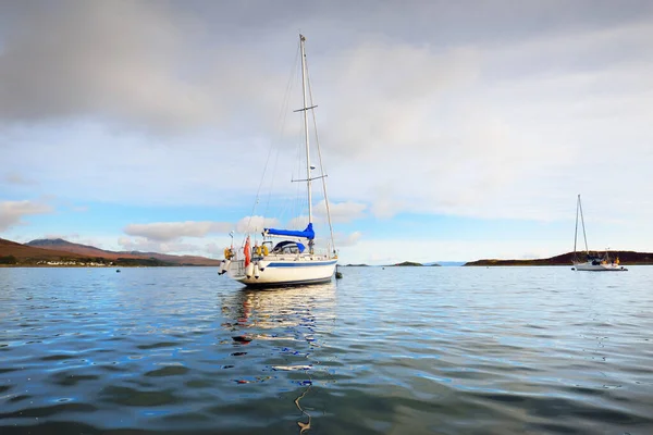 White sloop rigged yacht anchored on mooring near the shore of Craighouse at sunset. Mountains (Paps of Jura) in the background. Craighouse, Jura island, Inner Hebrides, Scotland, UK. Travel concepts