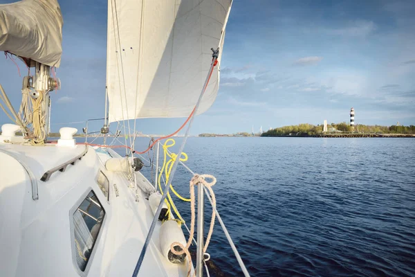 White yacht sailing in the Baltic sea after the rain at sunset. Riga, Latvia. Close-up view from the deck to the bow, mast and sails. Lighthouse in the background. Sport, recreation, travel