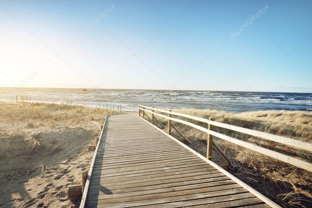 A wooden pathway to the Baltic sea at sunset. Sand dunes and plants (dune grass, Ammphila) in the background. Clear blue evening sky. Idyllic seascape. Latvia. Eco tourism, environment, ecology.