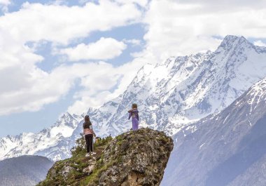 Kayanın tepesindeki kızlar kar kaplı Kafkaslar 'ın arka planına karşı. Ingushetia Cumhuriyeti, Rusya. 15 Mayıs 2021