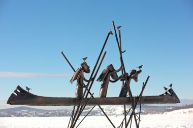 Nizhny Tagil, Russia - February 16, 2016: Metal sculpture of the three men, with wings on his back, which pluvut boat. The boat and the man sitting bird. The sculpture stands on the frozen pond. In the background - a mountain with a tower. clipart