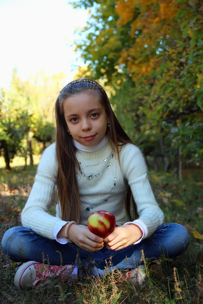 Flicka med långt hår som sitter i en park på marken med ett äpple i handen — Stockfoto