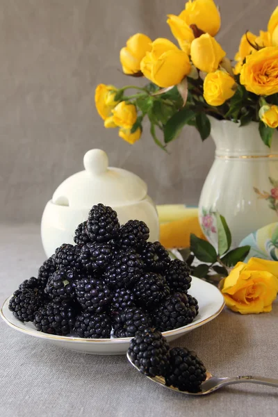 Saucer with blackberries and a vase with yellow roses — Stock Photo, Image